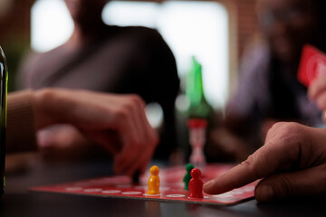 Obraz premium Close up of people hands moving plastic figurines on game table while sitting at table. Group of friends playing boardgames together while enjoying snacks and cold beverages.