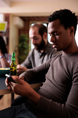 African american man with modern touchscreen smartphone sitting at table while browsing internet. Person sitting at home in living room while navigating on browser and social media feed.