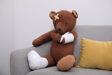 Toy bear with bandages sitting on sofa near light wall