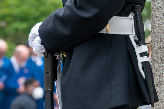 A Young Male Military Officer Stands At Attention Wearing A Dark Green Uniform With A Wide White Belt, Baton, And White Gloves. The Soldier Is Holding A Rifle. Veterans Are Standing In The Background.