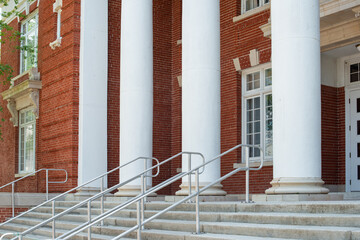 Four large white round columns at the facade entrance to a red vintage brick building. There are multiple panes of glass in tall white windows. The limestone steps have stainless steel handrails.   