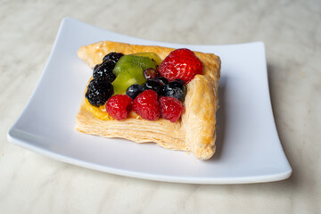 A square fruit filled French puff pastry on a white square plate. The cream and custard filled dessert have strawberries, kiwi, and blackberries on top with a sugar glaze. The pastry is flaky.	