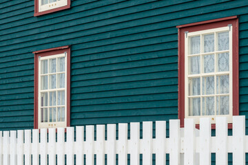 A vibrant teal green colored exterior wooden clapboard wall of a country style house with a white picket fence. There are multiple glass double hung windows with red trim and lace curtains hanging. 