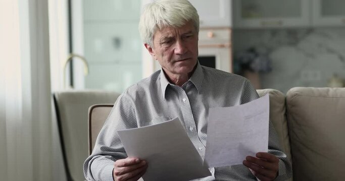Serious senior older grey haired man reading paper documents. Freelance businessman reviewing reports, receiving good news, getting happy, smiling, sitting on sofa at home