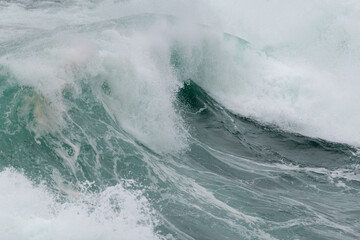 An angry turquoise green color massive rip curl of a wave as it barrels rolls along the ocean. The white mist and froth from the wave are foamy and fluffy. The ocean in the background is deep blue. 