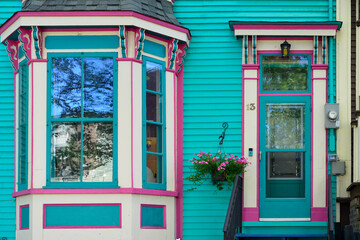Fototapeta na wymiar The entrance to a colorful teal green wooden building with pink and cream colored decorative trim. The façade single door has a basket of flowers, a mailbox, light fixtures and ornamental moldings. 
