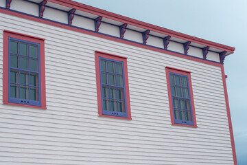 The roof section of a large white vintage wooden building with decorative pink and purple wood trim. There are three multi-pane windows on the top floor. The background is a dramatic blue cloudy sky.
