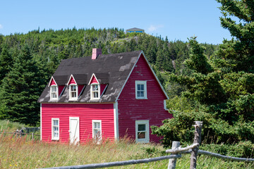 A view of a vintage cottage under blue sky and clouds. The small red wooden building has multiple double hung windows with a black roof. There's an old log fence, green grass, and a gravel path. 