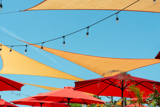 Multiple Triangle Shaped Yellow Nylon Sunshades And Awnings Hanging Over A Patio Deck. There Are Red Colored Canvas Umbrellas Hung With Strings Of Clear Patio Light Against A Bright Blue Sunny Sky.