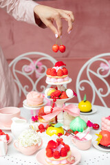 Vertical photo of cropped woman hand putting cherry on sweet desserts on cake stand on table. Open of new cafe or bakery