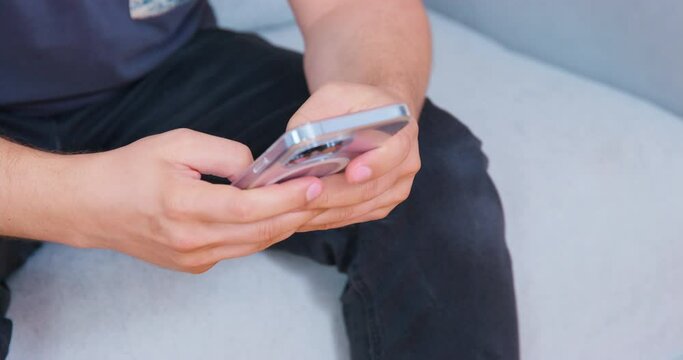 Man scrolls through social media news sitting on light grey mild sofa. Hands hold silver coloured modern mobile phone. Guy wears casual clothes