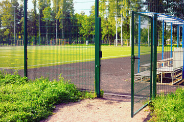 A large open metal door in a painted metal mesh fence, behind which you can see a green football...