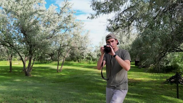 Photographer taking pictures in direction of camera using camera DSLR with zoom lense and wearing baseball cap with green nature background