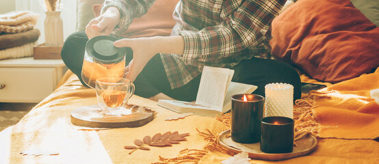 Woman's hand pouring tea in a cup while sitting on bed, reading book, relaxing at home in autumn fall season. Concept of cozy autumn at home, hygge. Long banner - 525449321