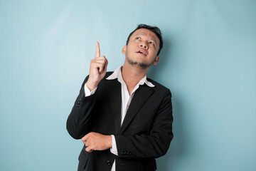Excited Asian man wearing suit pointing at the copy space upside him, isolated by blue background