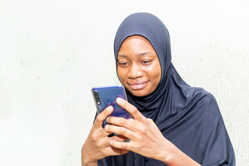 excited young Nigerian muslim woman dressed in hijab using mobile phone chatting online