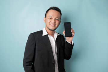 A portrait of a happy Asian businessman is smiling and holding his smartphone showing copy space on it's screen wearing black suit isolated by a blue background