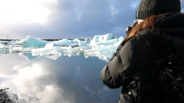 cinematic shot of a woman taking photos of the landscape and the icebergs found in the Jökulsárlón national park in Iceland.