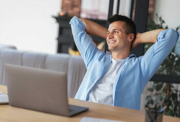 Young male businessman satisfied with the completed project sitting with their hands behind their heads relaxing at work in the office, taking break after long work at the laptop