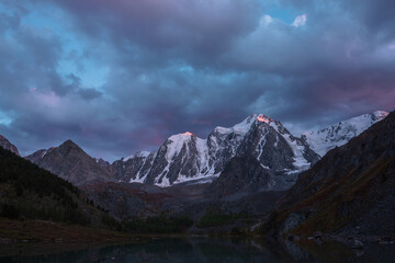 Obraz premium Atmospheric autumn twilight landscape with sunset gold reflection on huge snowy mountain top in violet dramatic sky. Mountain lake with view to giant snow-covered mountains under cloudy sky in dusk.