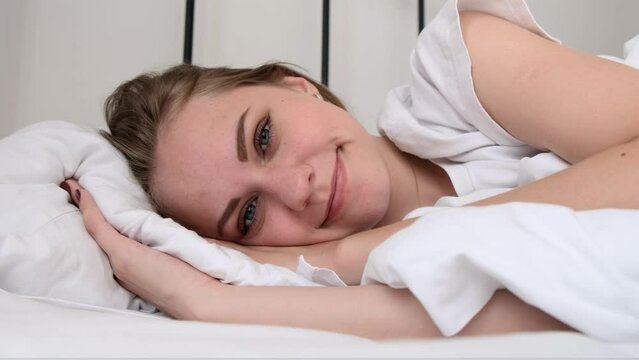 Natural beauty. Young seductive smiling woman looking into camera lying on her side in bed. Attractive girl strokes bed with her hand and laughs, lying on pillow. Rest, recovery. Slow motion, 4k
