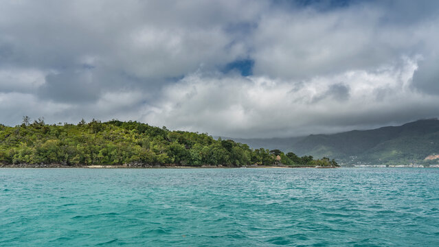 The tropical island is completely overgrown with green vegetation. Turquoise ocean and white cumulus clouds in the sky. Seychelles