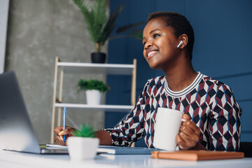Young African American woman entrepreneur having a call, sitting at her office desk and drinking...