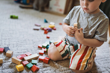 A little boy, 2 years old, sits on the floor and carpet at home and plays with colored wooden blocks. Educational toy constructor. The child learns to play according to the Montessori method - Powered by Adobe