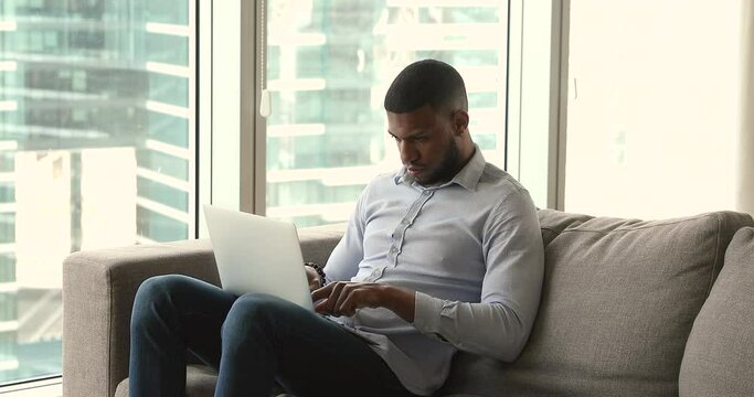 Handsome African man sit on sofa, using modern wireless computer, working in modern skyscraper flat, typing e-mail on laptop keyboard looks focused. Workflow, lifestyle, business, technology concept