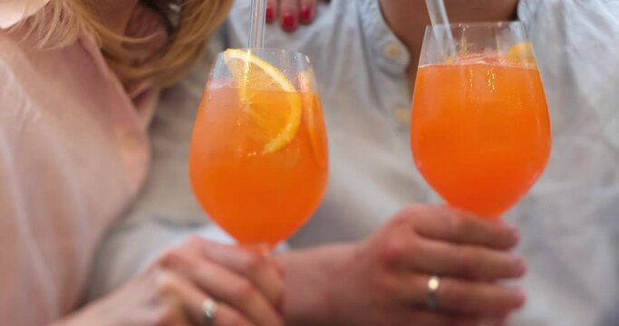 Romantic couple clinking with glasses of Spritz in bar. Aperol spritz cocktail, close up shot. Refreshing alcoholic drink in the hands. No faces. High quality 4k footage