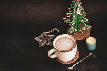 Obraz na płótnie Canvas Coffee with milk in a light ceramic mug, a wooden figure of a Christmas tree and a coffee spoon on a very dark background. Composition with New Year's decor