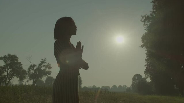 Silhouette of elegant attractive African American woman in sundress holding hands in prayer, praying for god bless and mercy while standing on green field over beautiful summer sunrise.