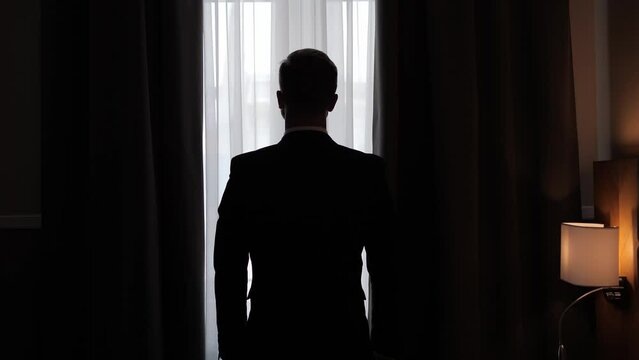 A successful businessman in a formal suit opens the curtains in a hotel room during a business trip in the morning. Business man silhouetted shot from back. Confident male CEO top manager new day.