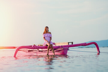 Naklejka premium Walk on the boat. A beautiful young woman in a striped sundress poses sitting on a boat with her feet in the water. The horizon line is in the background. Travel and recreation