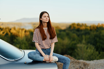 A woman car driver sits on the trunk of a car and looks into the distance admiring a beautiful view...