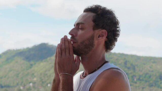 Peaceful bearded man quietly says the prayer, looks upward at beautiful sky, with hope while praying to God with his eyes closed, standing lonely in the beautiful nature with huge mountains background