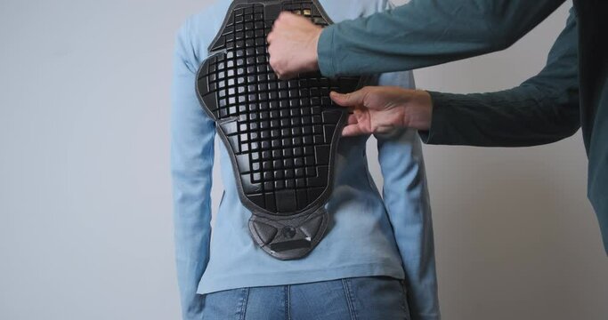 Man tries on a plastic back protector for a motorcycle jacket on a woman standing with her back. He taps his hand on the shield that covers his back.