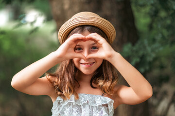 Portrait of a little Caucasian girl in a straw hat showing a heart sign with her fingers. There is...