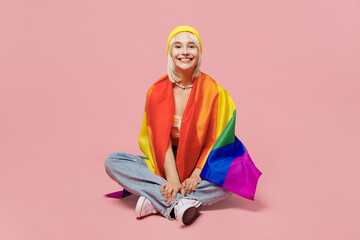 Full body young happy blond lesbian woman wrapped in flag 20s she wear yellow hat sitting on floor look camera isolated on plain pastel light pink background. People lgbtq lifestyle fashion concept.