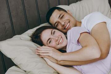 Young smiling happy fun calm lovely caucasian couple two family man woman she he wearing t-shirt pajama lying in bed rest spending time together in bedroom home in own room hotel. Real estate concept.