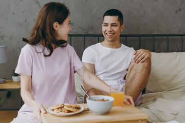 Young smiling happy fun couple two family man woman she he wear t-shirt pajama eat breakfast sitting in bed rest spend time together on weekends in bedroom home in own room hotel. Real estate concept.