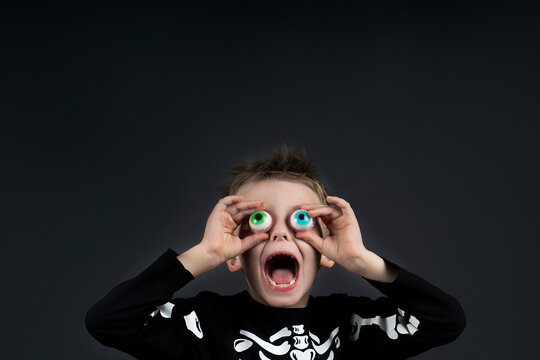 Cute little halloween boy with toy eyes looking  up and open mouth with shocked and delight copy space wearing Halloween costume skeleton on dark background with space for text . Space for advertising