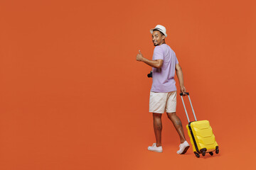 Full body traveler black man in purple t-shirt hat go with suitcase show thumb up isolated on plain orange background Tourist travel abroad in spare time rest getaway Air flight trip journey concept