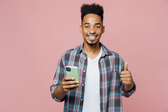 Young smiling happy fun man of African American ethnicity 20s wear blue shirt hold in hand use mobile cell phone show thum up isolated on plain pastel light pink background. People lifestyle concept.