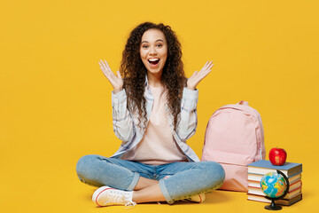 Full body young black teen girl student she wear casual clothes backpack bag sitting on floor near books spread arms isolated on plain yellow color background High school university college concept.