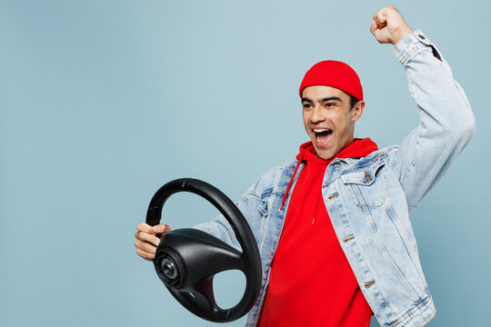 Young middle eastern man 20s he wear denim jacket red hat hold driving car hold steering wheel do winner gesture isolated on plain pastel light blue cyan background studio. People lifestyle concept.