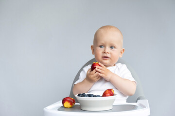 Baby boy in white t-shirt eats organic fruits and berries on grey background. Healthy fruit snack - peach, blueberries, healthy diet, feeding, solid food. Feeding chair. Vegan lifestyle concept
