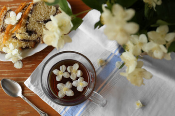 Glass cup of aromatic tea with beautiful jasmine flowers and dessert on wooden table, flat lay