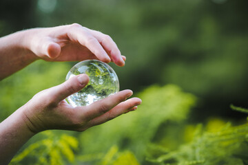 hands holding transparent glass ball or  crystal who reflects trees of a forest outside in green nature sphere symbolizing save the earth or planet