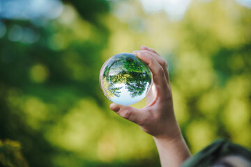 hands holding transparent glass ball or  crystal who reflects trees of a forest outside in green...